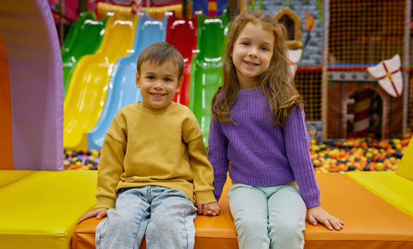 two children sitting on colorful play area seating in a fun indoor playground with slides and balls featuring vibrant colors and playful atmosphere 7 fun activities 7 playful moments 7 smiles