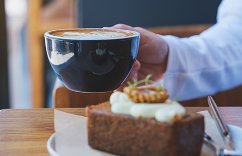 a person holding a black coffee cup with latte art beside a slice of cake with cream and decorative elements enjoying a cafe moment with 2 treats